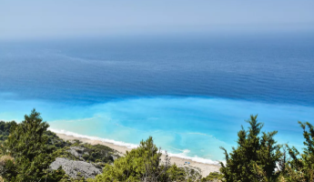 Blick auf das t&uuml;rkisfarbene Meer und den Strand von Samos, eingerahmt von gr&uuml;nen B&auml;umen.