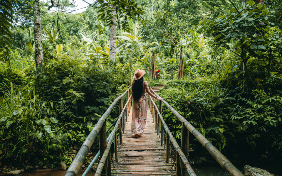 Die junge Frau in Kleid und Hut geht &uuml;ber die Br&uuml;cke des Wanderwegs zum Leke-Leke-Wasserfall und blickt sich in einem tropischen Wald um